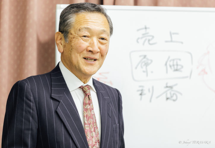 経営の考え方を穏やかな表情で語る講師と、背後に理念を書いたホワイトボードの様子