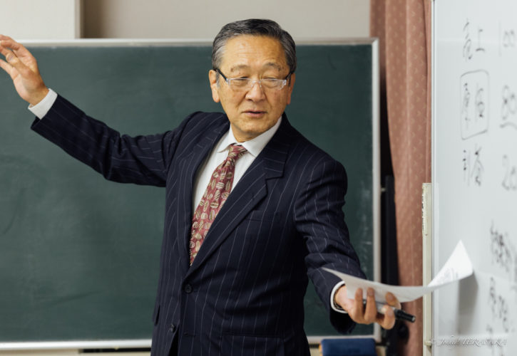 度量と器量をテーマに、経営者の品性について語る講師が、黒板の前で資料を手に説明している講義風景