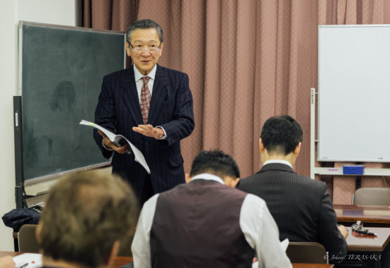 講義室で資料を手に、受講者に向けて穏やかに語りかける講師の姿