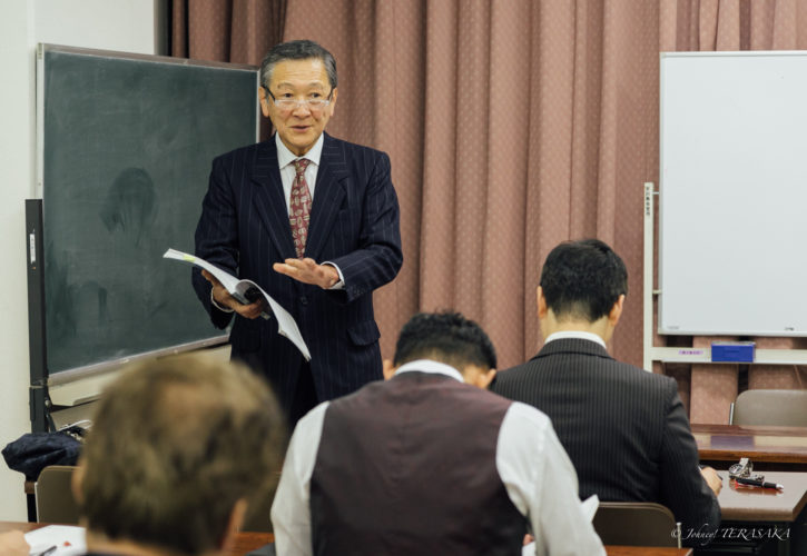 講義室で資料を手に、受講者に向けて穏やかに語りかける講師の姿