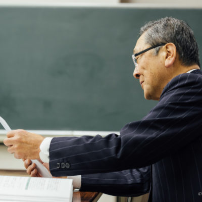 講義室で資料を手にし、黒板の前で内容を確認しながら静かに語りかける講師の横顔