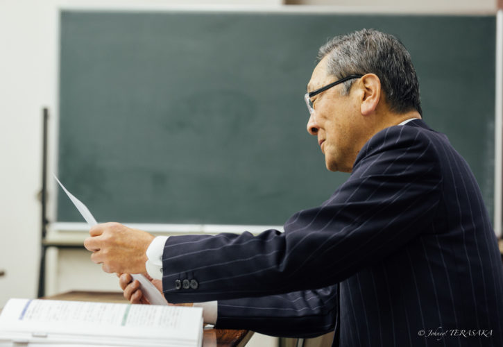 講義室で資料を手にし、黒板の前で内容を確認しながら静かに語りかける講師の横顔