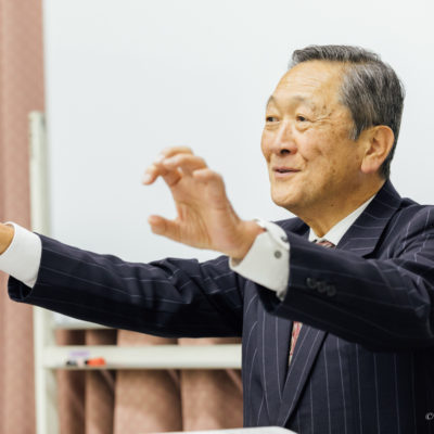 経営の本質について両手を広げながら語りかける講師の姿。参加者に向けて全体像と流れを伝えている講義風景。