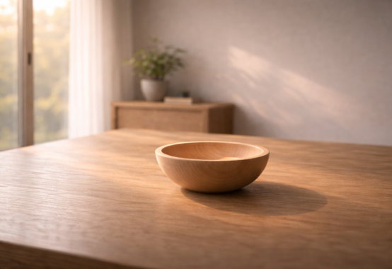 柔らかな自然光が差し込む木の机の上に置かれた木の器と、周囲の静かな余白。人を受け入れ周囲を活かす「器」の思想を象徴する静かな情景。
